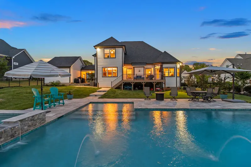 Pool glowing at twilight behind a two-story home with lit windows